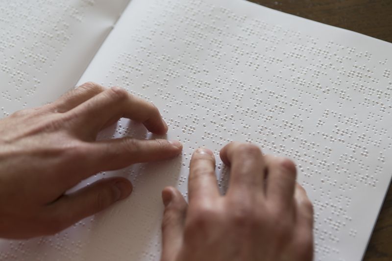 Zwei Hände ertasten die Buchstaben auf einer Seite, die mit Braille bedruckt ist.