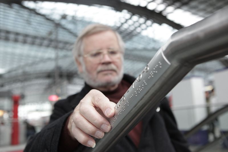 Ein Mann tastet mit der rechten Hand nach der Braille-Markierung an einem Treppengeländer.